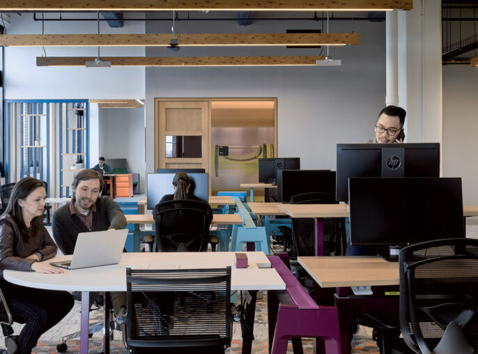 Modern open-plan office space featuring eclectic furniture with a combination of communal desks, private booths, and a lounge area, accented by vibrant carpets and exposed ceiling beams. Employees are engaged in collaborative and individual work under ample natural and ambient lighting.