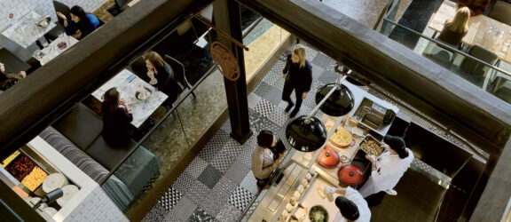 Overhead view of a bustling restaurant interior showing a contrast of textures with exposed brick walls, sleek wooden beams, geometric tiled flooring, and an eclectic mix of modern and rustic furniture; diners enjoy meals under warm, ambient lighting.