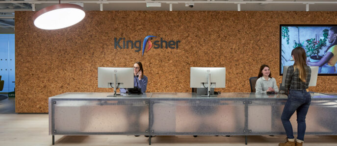 Modern office reception area with a warm-toned wooden accent wall featuring a company logo, flanked by sleek, minimalist workstations and punctuated by a large, oval pendant light. Staff engage with visitors against the backdrop of a contemporary digital display.