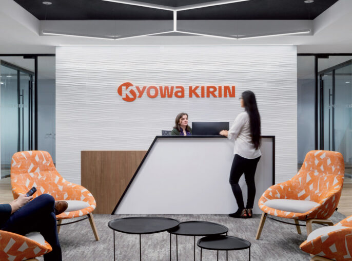 Modern corporate reception area featuring bold Kyowa Kirin brand wall, pops of orange in the upholstered armchairs, sleek black center tables, and dynamic lighting overhead, creating a welcoming ambiance for professionals and guests.