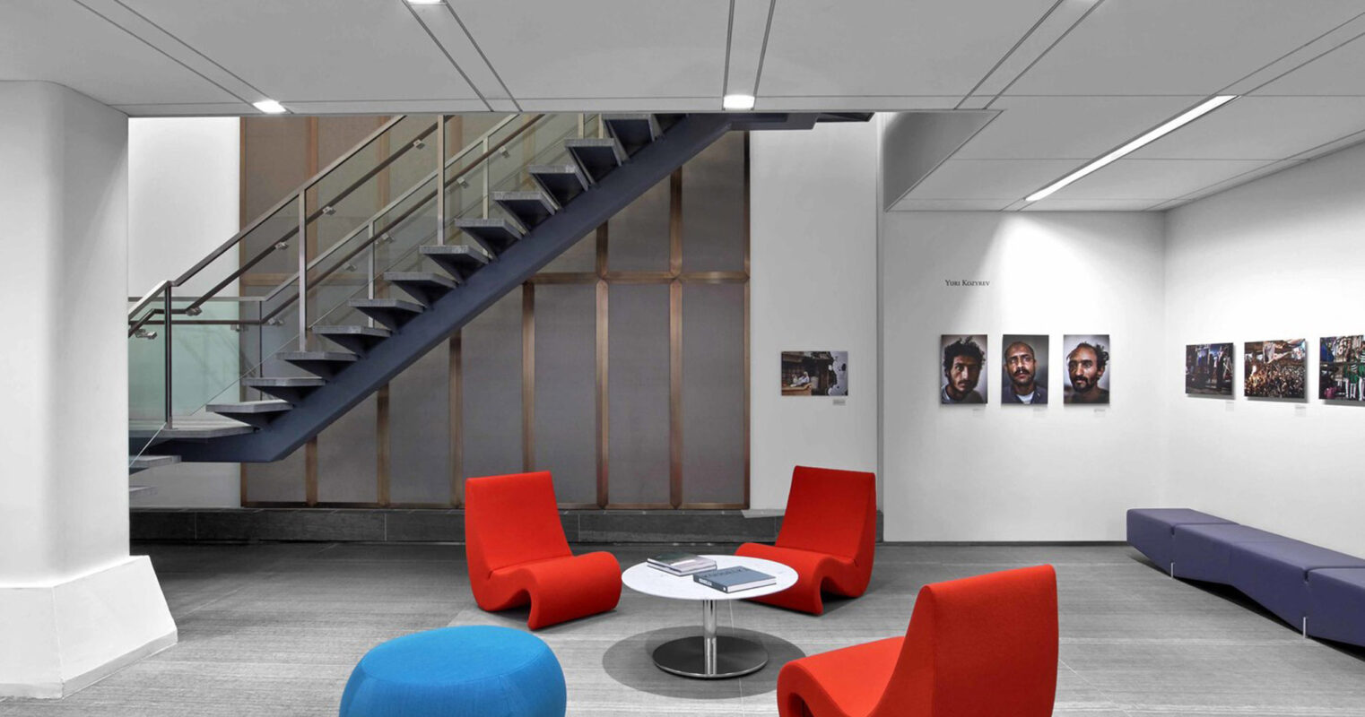 Modern interior with a minimalist staircase featuring glass balustrades, alongside a vibrant seating area with red armchairs, blue ottomans, and an elongated purple bench. The neutral palette is accentuated by strategic lighting and framed artwork on the walls.