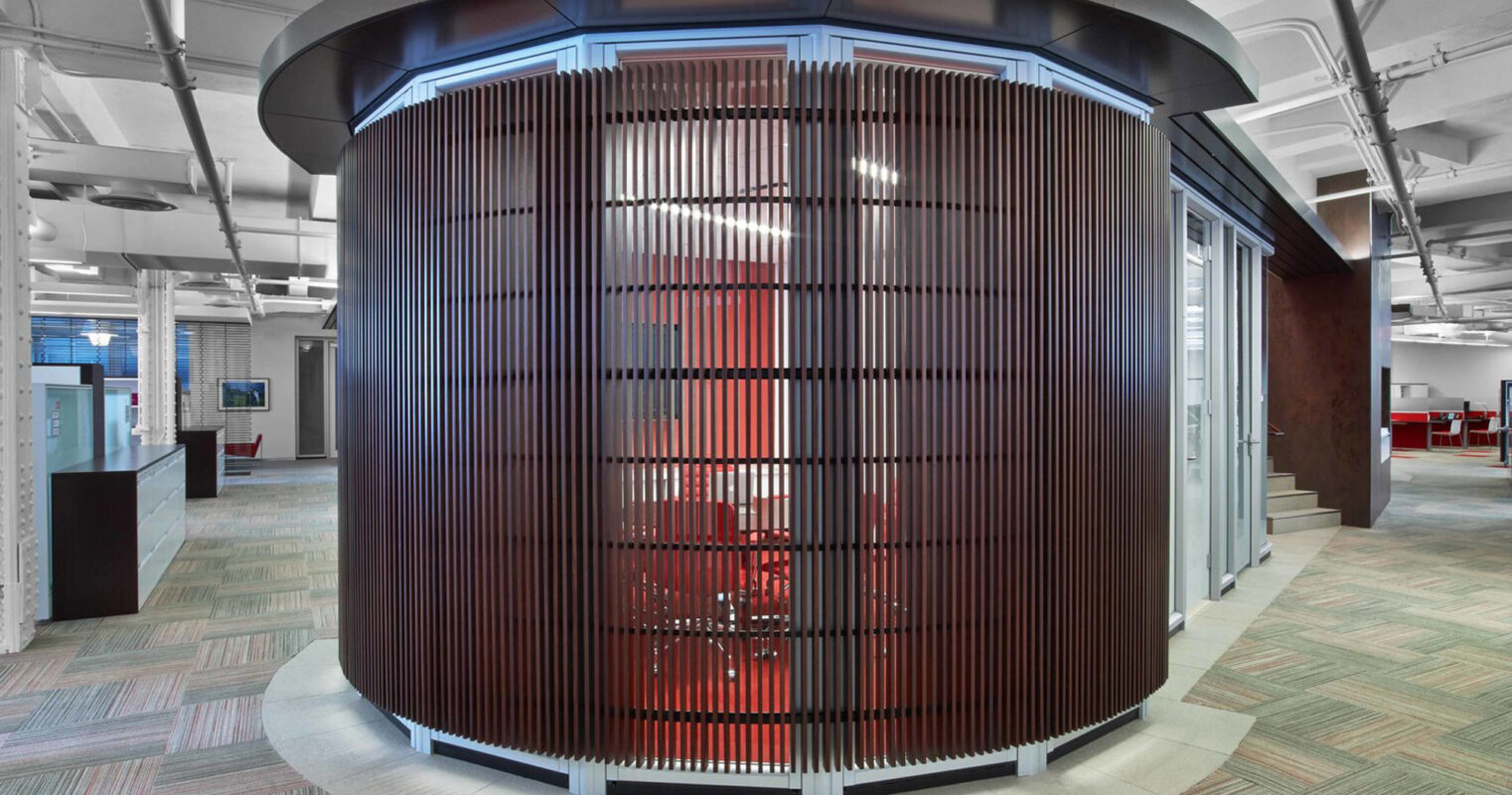 Modern office space featuring a cylindrical core with vertical wooden slats, surrounded by diverse seating areas. Above, a blue-tinted circular light fixture echoes the core's form, contrasted against exposed white beams and ductwork. The floor combines a palette of geometric-patterned carpet tiles.