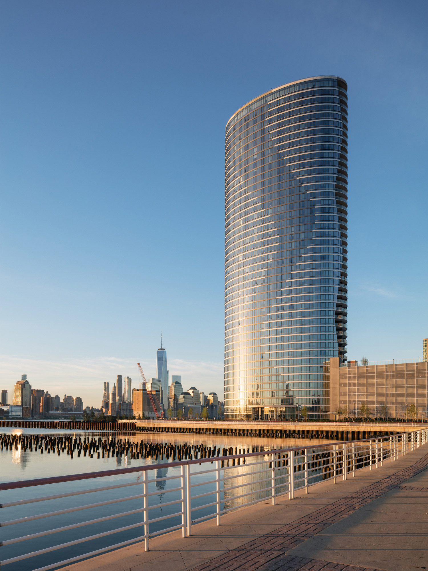 Modern high-rise building with curved glass facade reflecting the sunset's warm hues, featuring a sleek, cylindrical architecture that stands out against a clear blue sky with a city skyline backdrop.