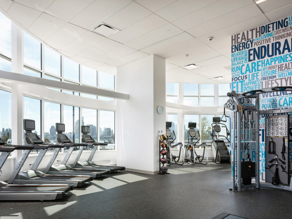 Modern gym interior with floor-to-ceiling windows providing abundant natural light, highlighting a row of treadmills and various exercise machines. The room features motivational words on a graphic accent wall, enhancing the energetic atmosphere.