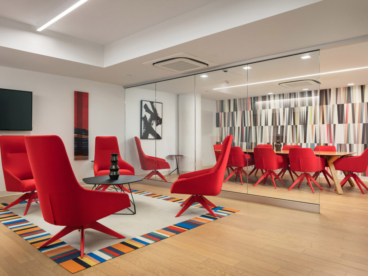 Modern office lounge with vibrant red chairs, sleek white surface tables, and a colorful striped accent wall. Overhead, contemporary rectangular lighting fixtures complement the room's bright, inviting ambiance.
