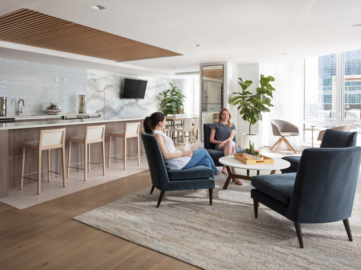 Open-concept living area with light wooden floors and white walls, featuring a kitchen with bar seating, modern appliances, and a lounge area with stylish navy armchairs and a plush sofa. Large windows afford city views, complemented by a serene color palette and contemporary artwork.