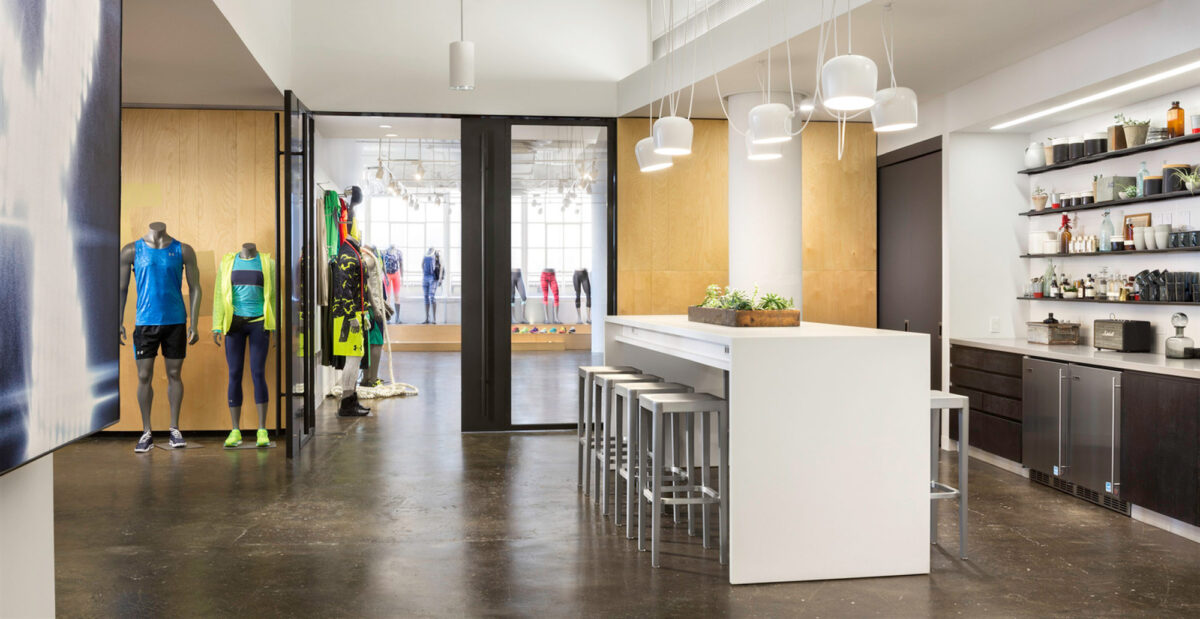 Spacious retail interior showcasing a blend of natural wood and modern materials, featuring pendant lighting, a central bar with stools, and striking displays of athletic apparel.