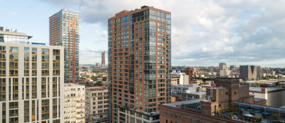 Modern urban landscape showcasing a mix of high-rise residential buildings with varied architectural styles, including glass facades and terracotta-colored brickwork, under a dynamic sky with scattered clouds.
