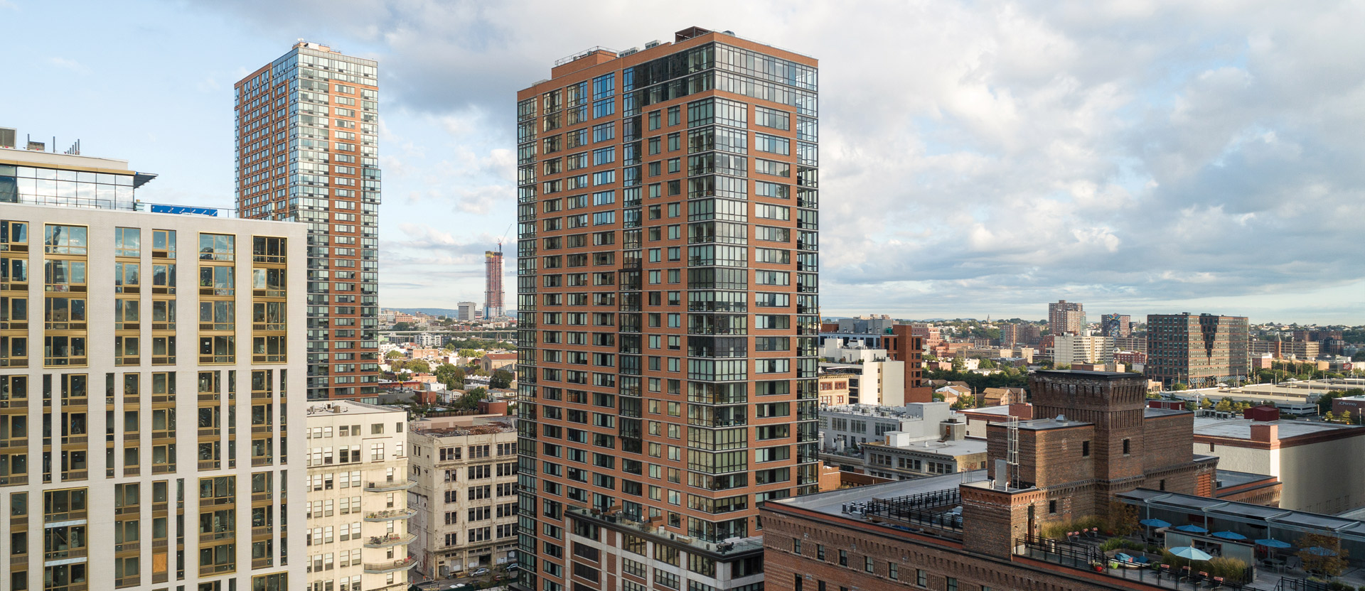Modern urban landscape showcasing a mix of high-rise residential buildings with varied architectural styles, including glass facades and terracotta-colored brickwork, under a dynamic sky with scattered clouds.