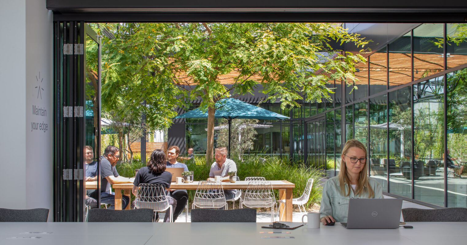 Professionals gather at a modern outdoor meeting space featuring minimalistic furniture and lush greenery, with a transparent glass facade integrating the interior office environment and the vibrant outdoor setting.