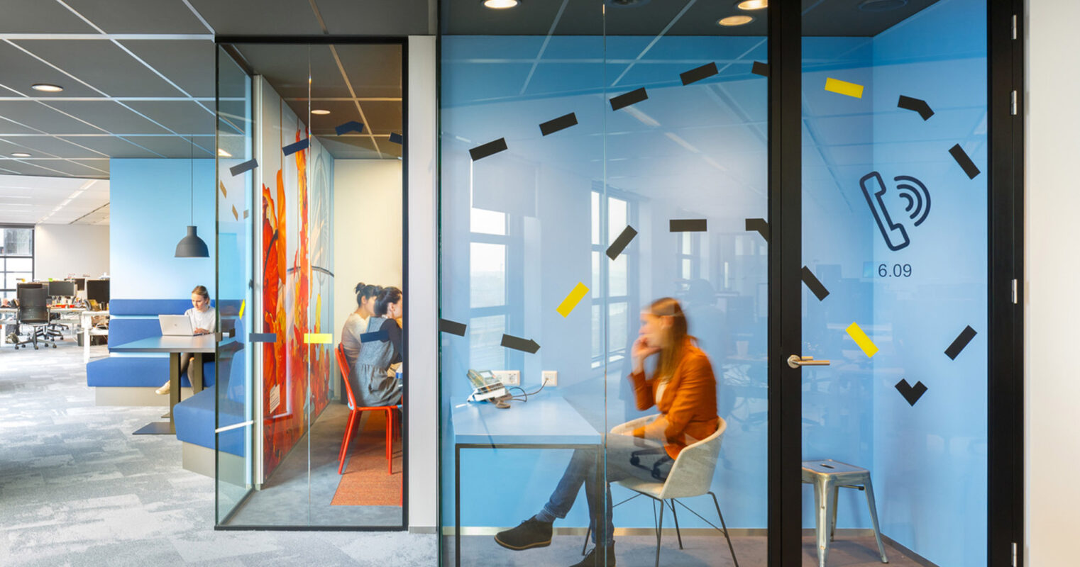 Modern office space featuring glass walls with dynamic geometric decals, fostering an open atmosphere while providing privacy. The vibrant contrasting carpet tiles and sleek furniture reflect contemporary design trends, encouraging a collaborative work environment.
