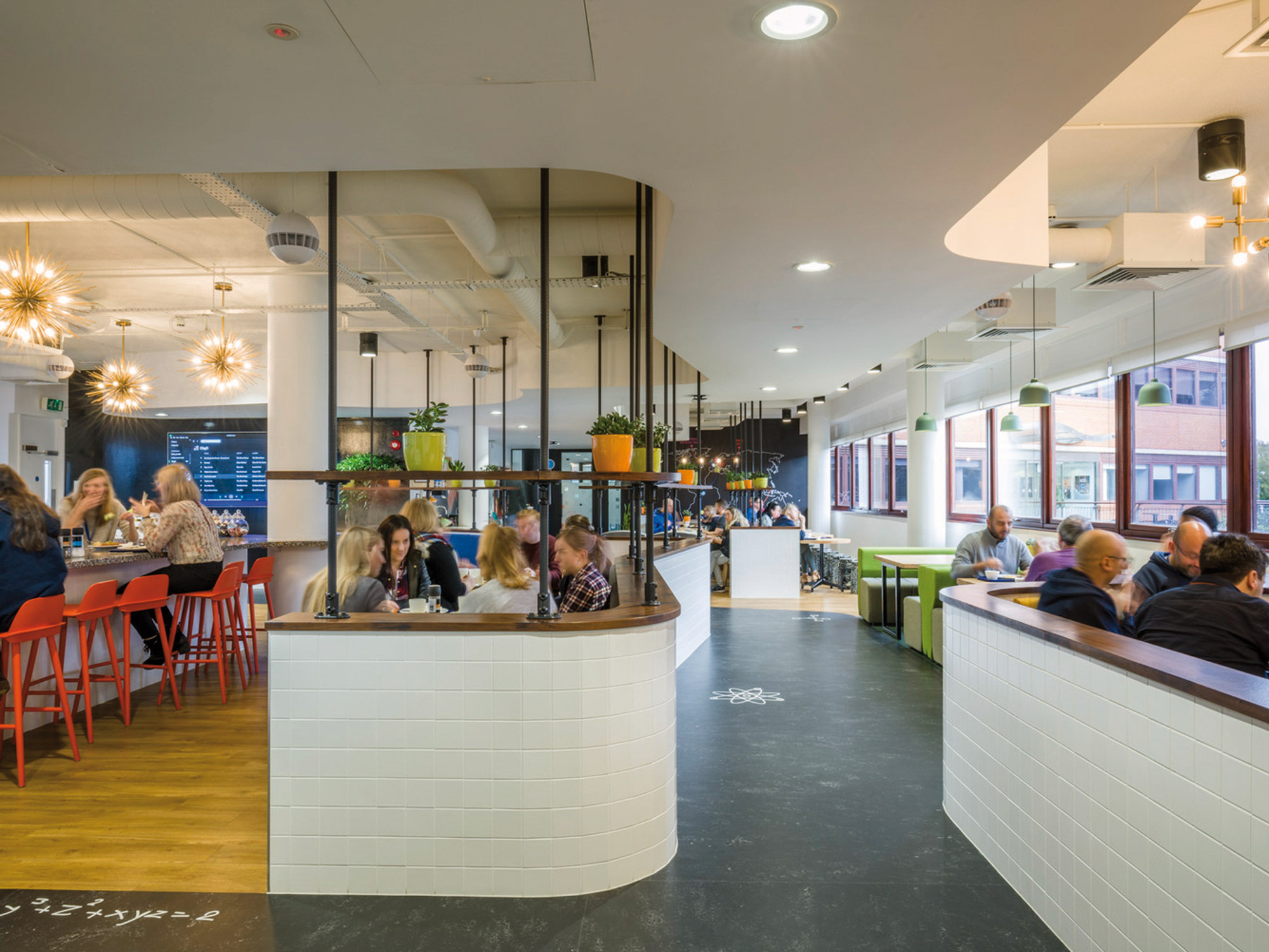 Modern cafeteria interior featuring sleek white counters, vibrant red and green seating. Overhead, unique starburst light fixtures illuminate the bustling, open-plan space with exposed ductwork, contributing to an industrial yet welcoming atmosphere.