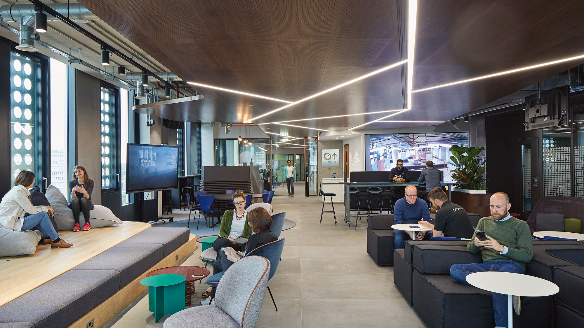 Modern office lounge area featuring tiered wooden seating, integrated LED lighting, and a mix of informal workspaces. The design encourages collaboration with circular seating accents and technological amenities, complemented by a palette of warm woods, greens, and neutral tones.
