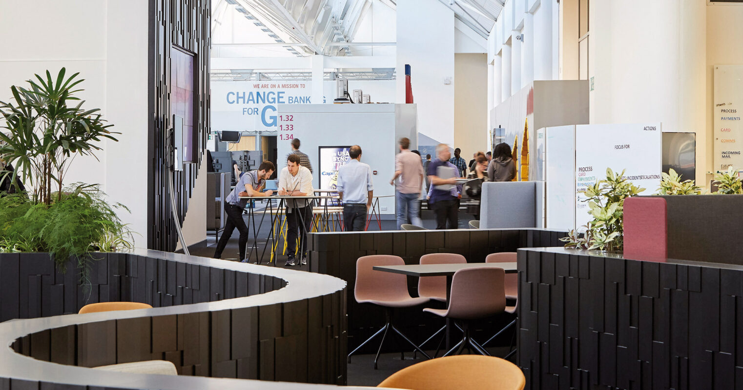 Modern exhibition space with geometric, intertwined seating arrangement featuring dark vertical slats. Overhead, natural light filters through a skylight, complementing the warm wooden flooring and the industrial-chic exposed ceiling beams. The gathering area is punctuated by round tables and earth-toned chairs, facilitating professional interaction.