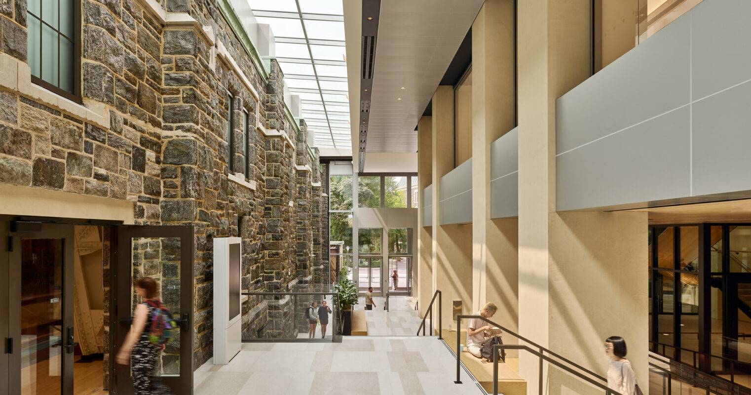 Spacious atrium with high ceilings, natural stone walls, and large floor-to-ceiling windows bathing the interior in natural light. The sleek metal balustrades contrast with the warm stone, enhancing the modern yet timeless aesthetic. White tiles in a grid pattern cover the floor, complementing the building's geometric lines.