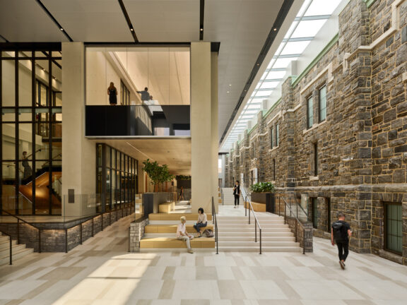Modern interior juxtaposing historical stone walls with glass partitions, sleek black metal frames, and a sunlit atrium. Elegantly crafted stairs and tiered seating areas facilitate flow and interaction, complemented by geometric floor patterns that enhance the space's contemporary yet timeless ambiance.