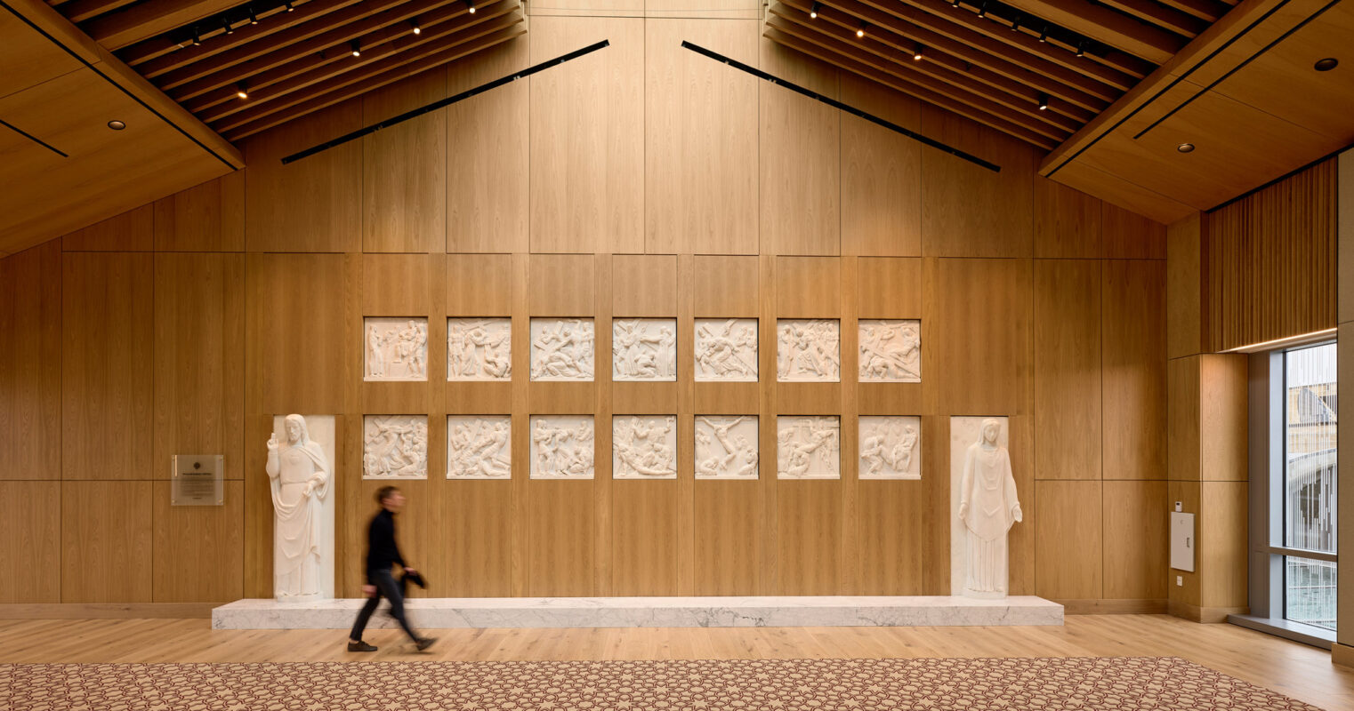 Modern gallery space boasting warm wooden panel walls and ceiling with geometric patterns, accentuated by ambient natural light filtering through a clerestory window. Elegant statues flank a series of framed artistic displays above a detailed patterned floor, with a person walking by, adding a dynamic element.