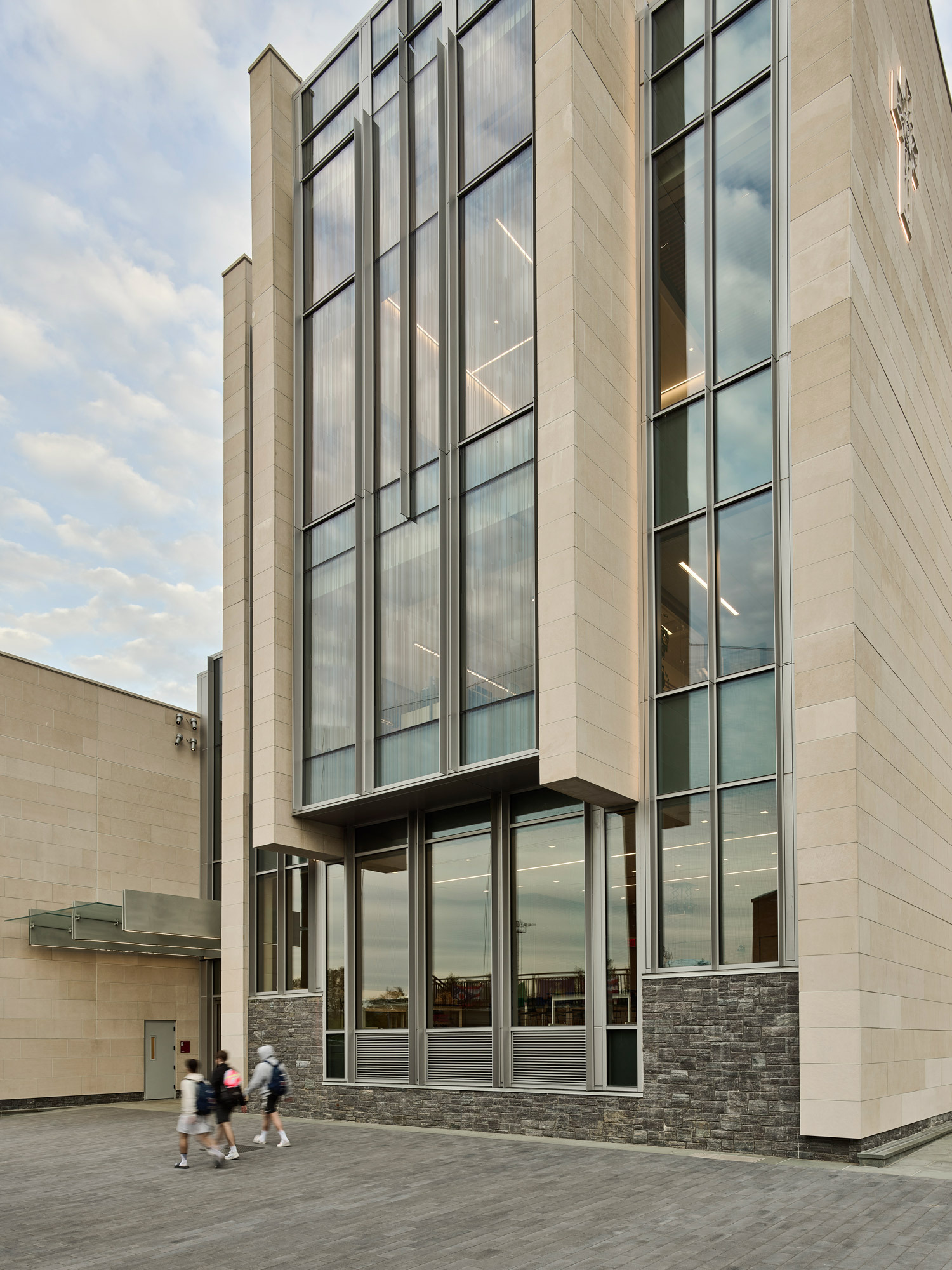 Modern glass-walled building with vertical and horizontal metal accents, featuring a rhythmic facade and an overhanging section for dynamic architectural interest, under a cloudy sky with pedestrians walking by.