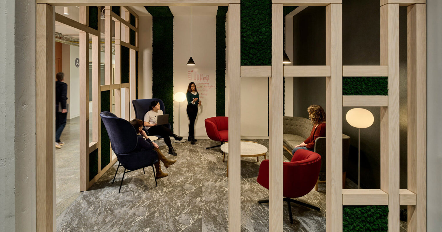 Modern office space with natural wood framing and green wall accents. Two individuals engage in work at tables with contemporary, dark blue armchairs under sleek hanging lights, enhancing the space's inviting ambiance.