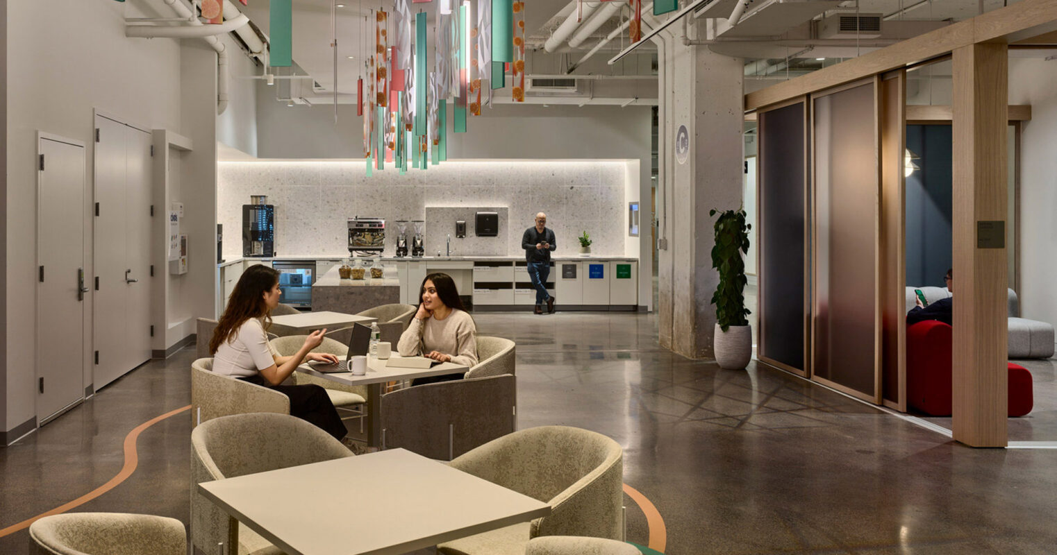Modern office lounge with hanging fabric art installation, neutral-toned furniture centered around a circular table, and a kitchenette in the background with a person standing by the counter. The open-concept area features concrete pillars and wood-trimmed glass partitions.