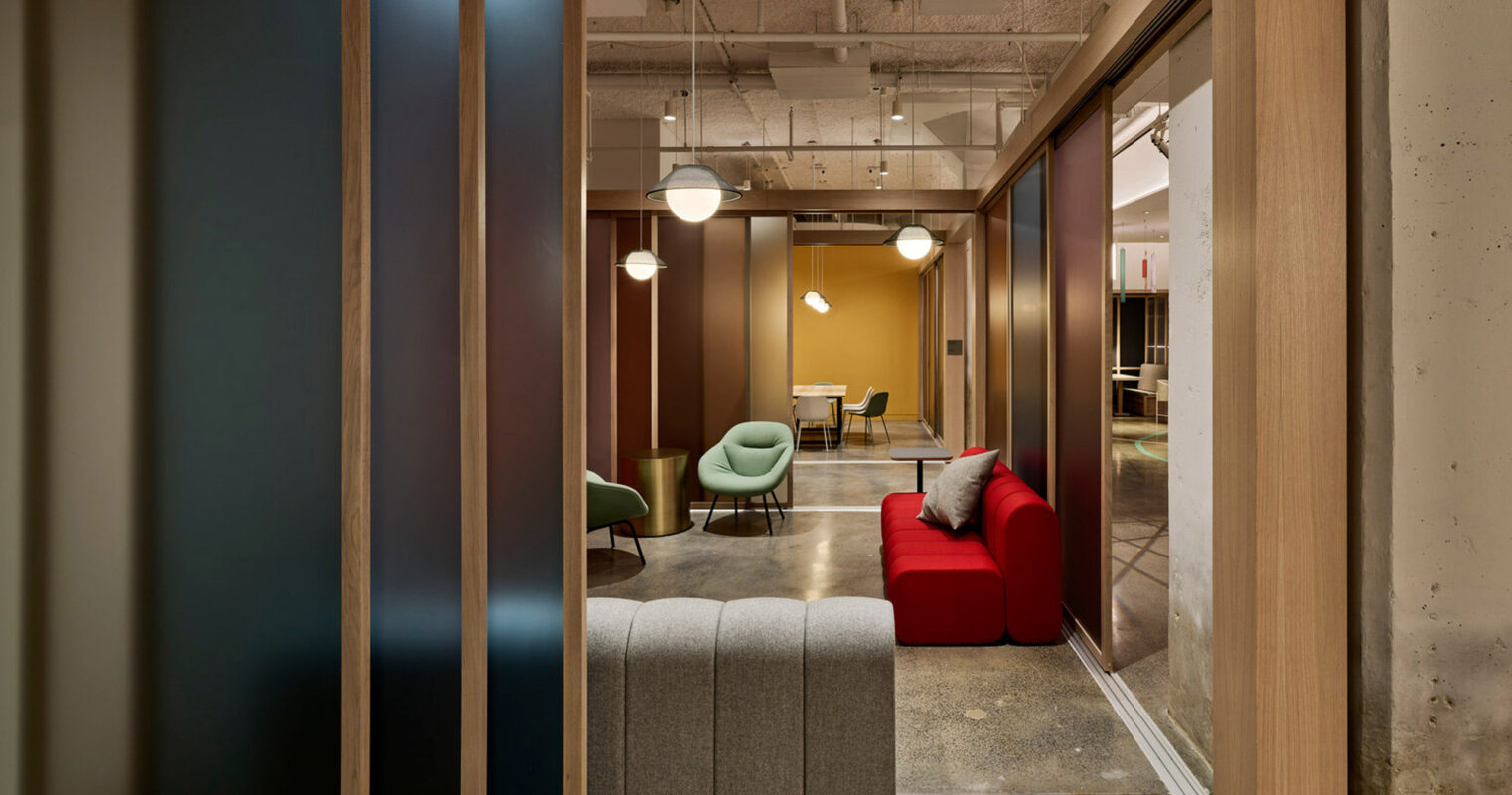 Modern office lounge with translucent sliding doors, featuring a mix of plush red armchairs and minimalist green chairs around a coffee table, complemented by warm pendant lighting and exposed concrete ceiling.
