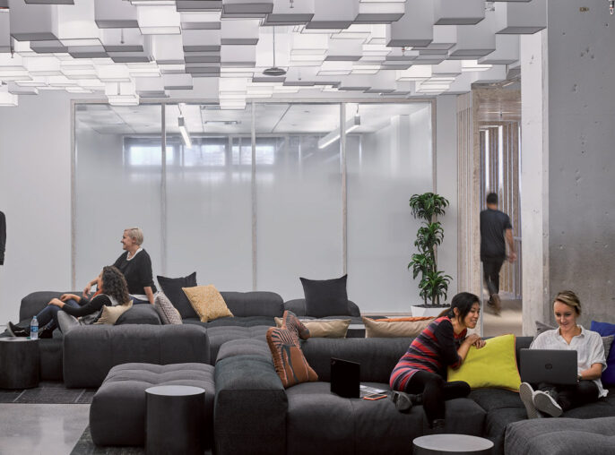 Modern office lounge with varied modular seating arrangements using plush, dark upholstered pieces. Overhead, a geometric pattern of rectangular light fixtures complements the exposed ceiling, creating a dynamic, sleek environment conducive to informal meetings or individual work.