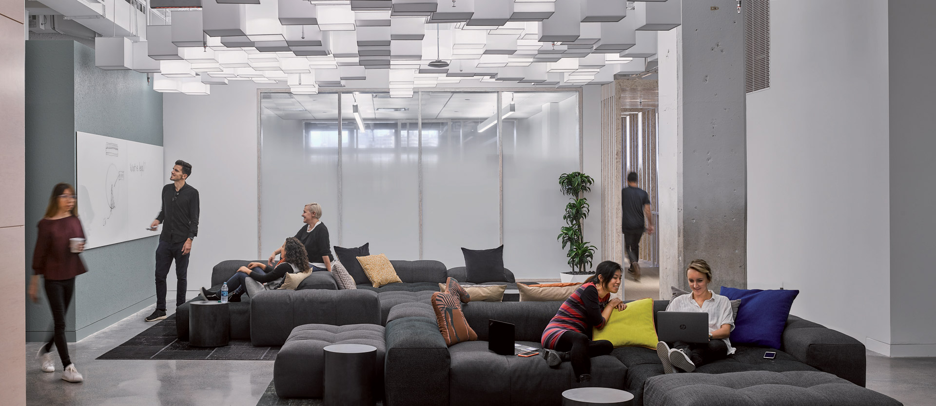 Modern office lounge with varied modular seating arrangements using plush, dark upholstered pieces. Overhead, a geometric pattern of rectangular light fixtures complements the exposed ceiling, creating a dynamic, sleek environment conducive to informal meetings or individual work.