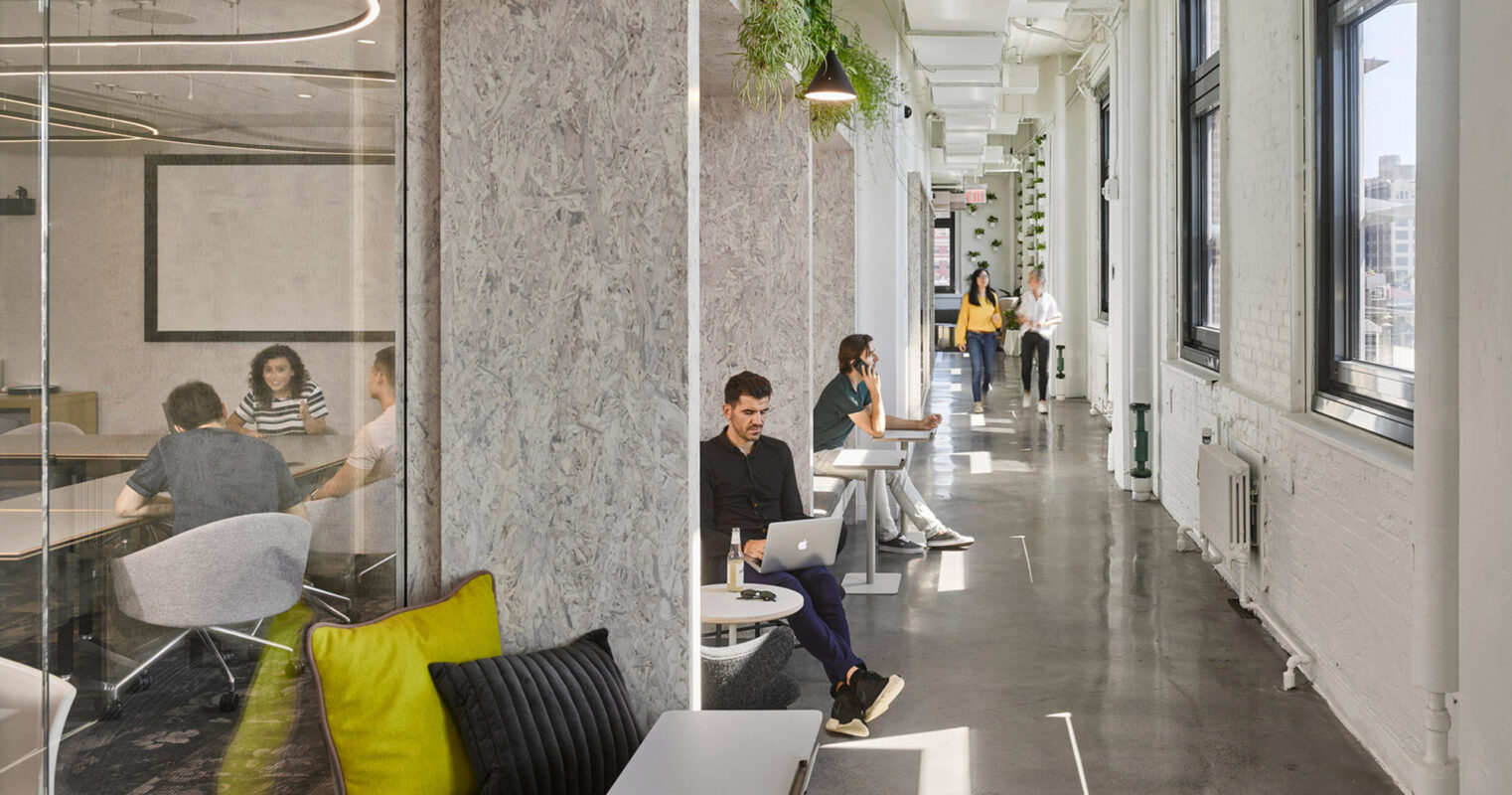 Modern office space blending natural light with greenery. Features a minimalist desk setup against marble walls, with employees working in communal zones that foster collaboration. Transparent partitions enhance openness, while plants add a biophilic touch to the sleek, contemporary environment.