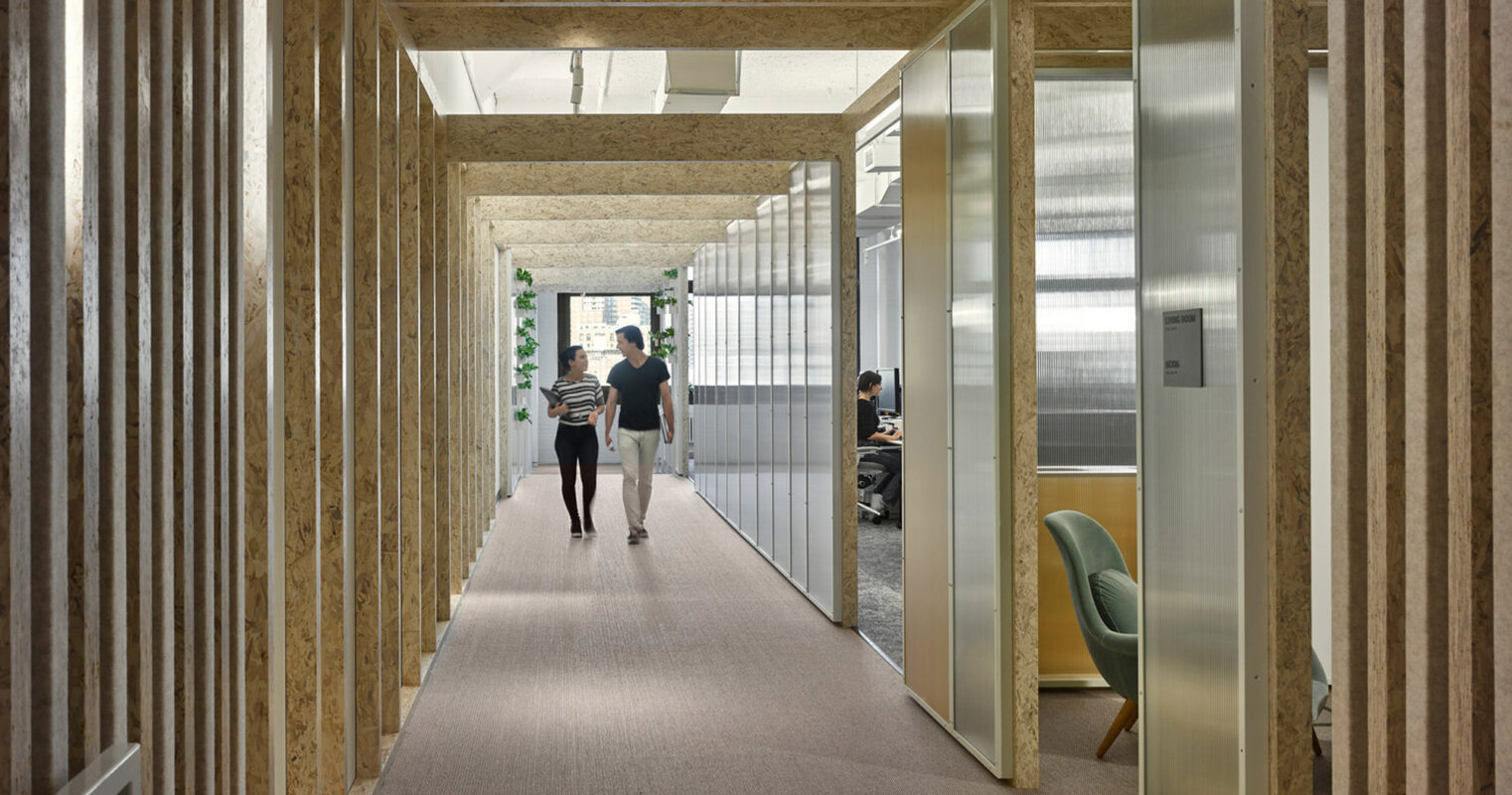 Narrow hallway in a modern office, lined with translucent glass and natural plywood walls, featuring a carpeted floor that leads towards a person in the distance. The space emphasizes natural light and transparency, fostering an open and airy work environment.