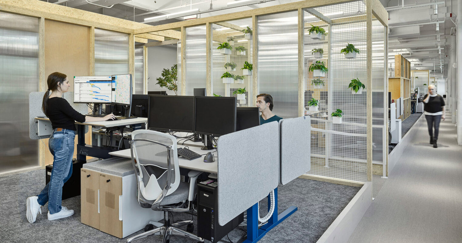 Modern office interior featuring standing desks, ergonomic chairs, and dual monitor setups. Glass partitions with wooden frames provide semi-private spaces, while indoor plants inject vitality. The workspace balances openness and privacy, optimizing collaboration and focus areas. Natural light accentuates the clean, contemporary aesthetic.