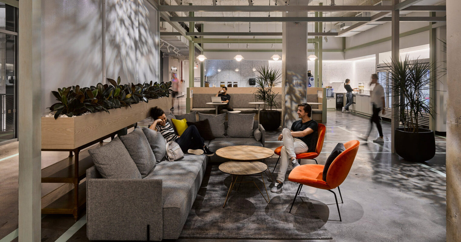 Contemporary open-plan office lounge with modular gray seating, round wooden tables, vibrant orange chairs, and wall-mounted planters. The space is enhanced by patterned natural light casting shadows, invoking a relaxed, collaborative atmosphere.
