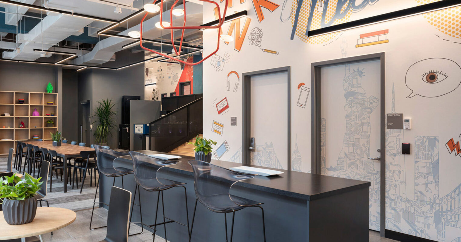 Modern office breakroom featuring a mural with playful typography, exposed ceiling with hanging red geometric light fixtures, sleek black countertop with high stools, and a communal wooden dining table.