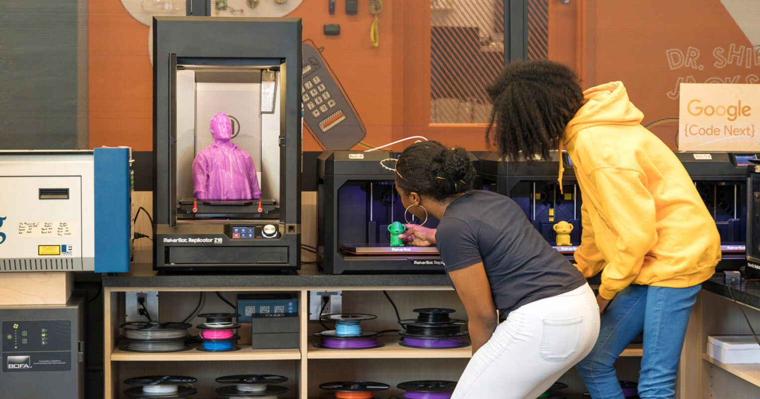 Two individuals observe a 3D printer in a modern maker space, featuring vibrant spools of filament on shelving and a range of prototyping equipment indicative of an innovative learning environment.
