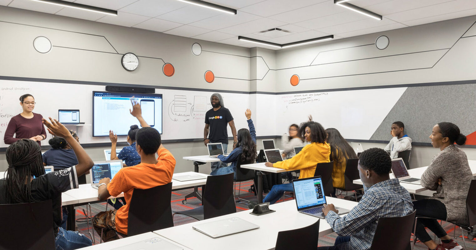 Modern classroom with vibrant orange and gray color scheme, featuring collaborative workstations, interactive whiteboard, and ceiling-mounted projector. Diverse group of students engaging in a dynamic learning session led by an instructor.