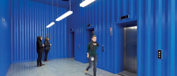 Vibrant blue corrugated metal walls contrast with a sleek white floor in a modern elevator lobby. Suspended linear lighting fixtures cast a well-distributed glow, while two individuals engage in conversation, adding a dynamic human element to the space.