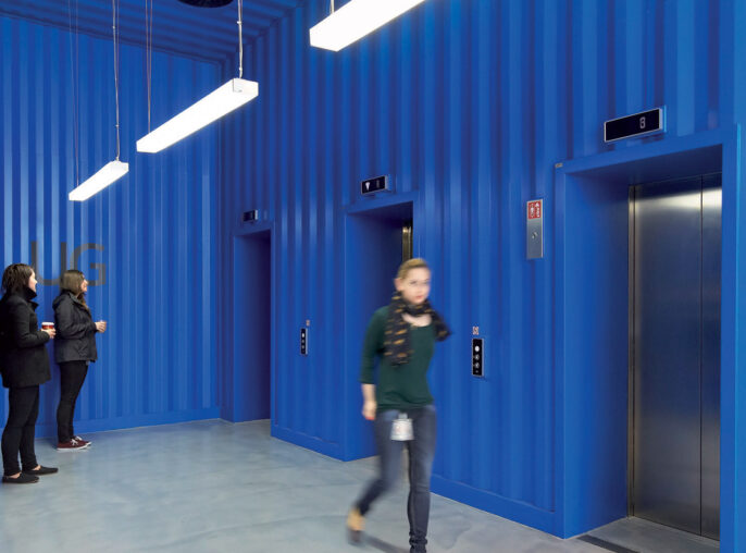 Vibrant blue corrugated metal walls contrast with a sleek white floor in a modern elevator lobby. Suspended linear lighting fixtures cast a well-distributed glow, while two individuals engage in conversation, adding a dynamic human element to the space.