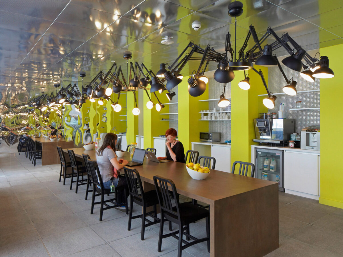 Modern cafeteria space featuring reflective ceiling with undulating pattern, industrial-style articulated black pendant lights, and communal wooden tables paired with minimalist stools. The vibrant lime accents contrast the muted tones, enhancing the space's energetic atmosphere.