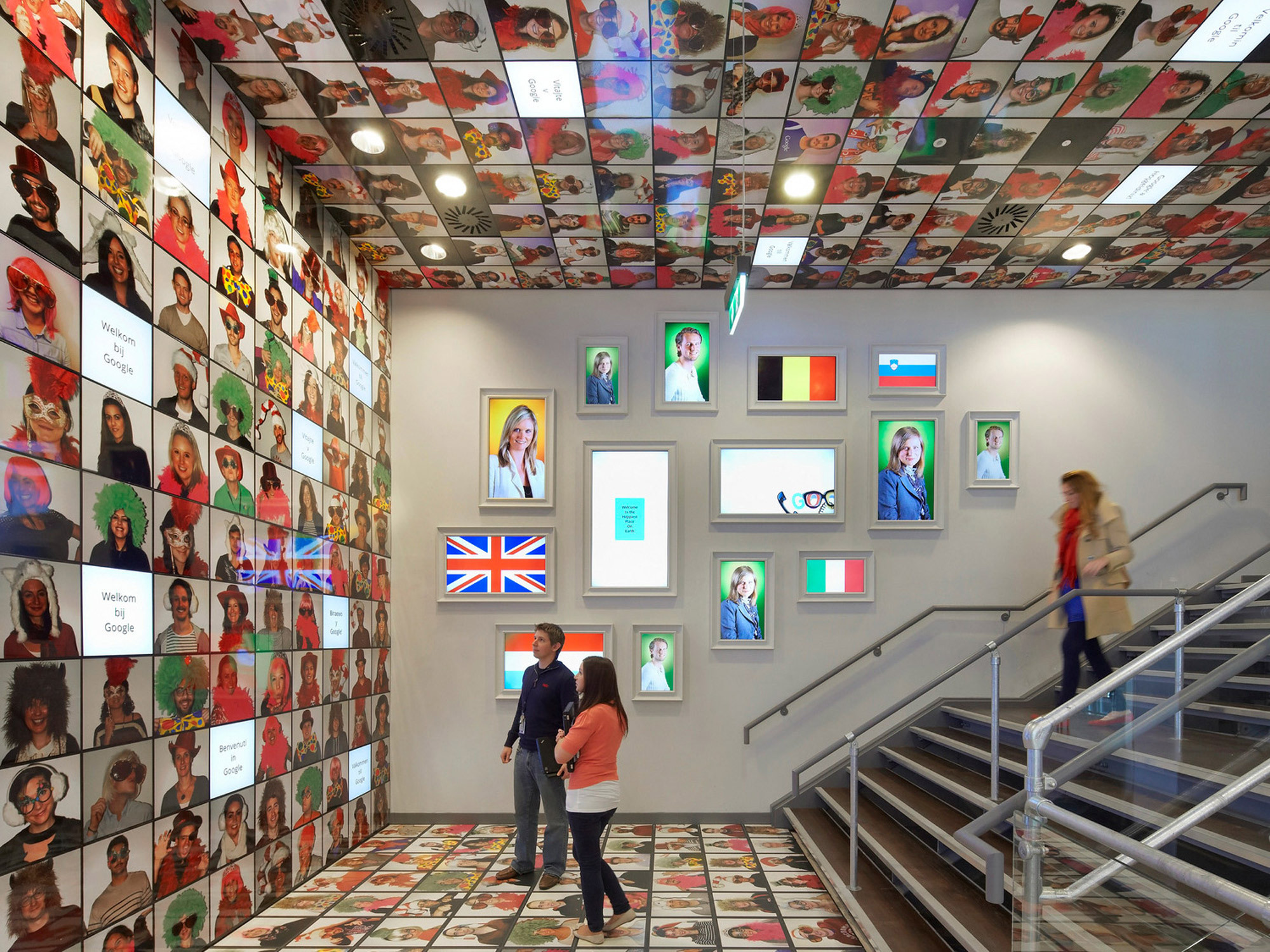 Vibrantly adorned staircase with a dynamic gallery wall, featuring portraits framed by a collage of multi-hued patterned wallpaper, which extends onto the ceiling, complemented by sleek metal railings and a woman ascending the stairs.