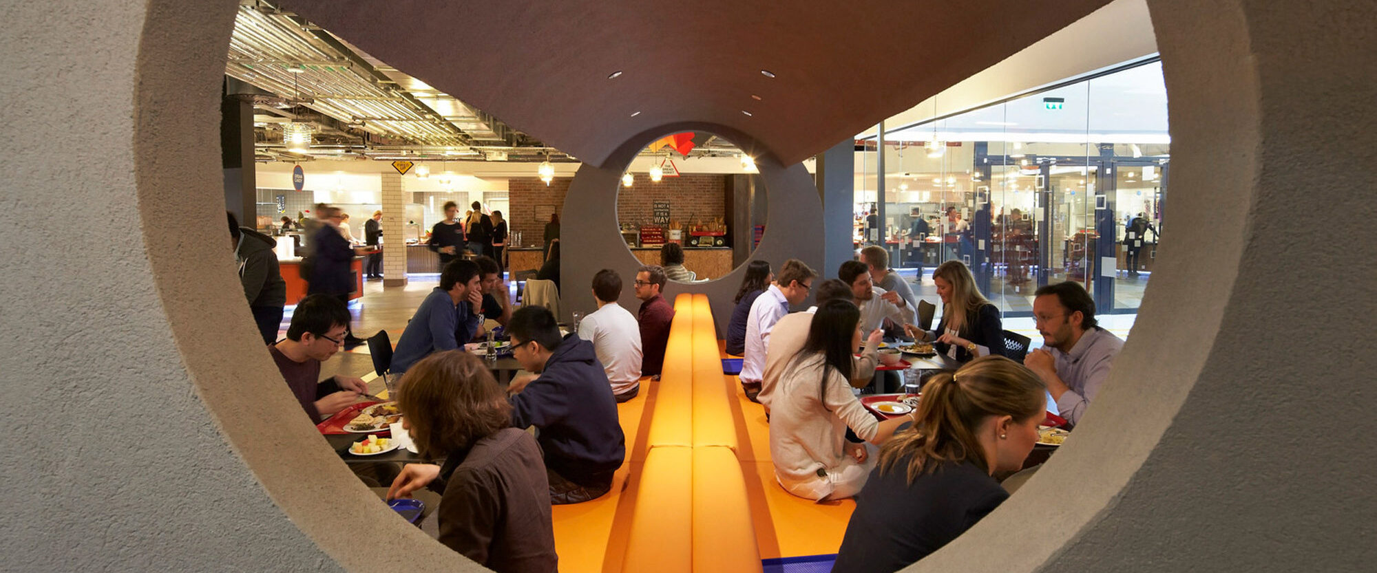 Circular framed view into a contemporary dining area, showcasing minimalist furniture, an orange carpet runway, and ambient lighting, amidst neutral-toned surroundings and diverse patronage engaged in conversation.