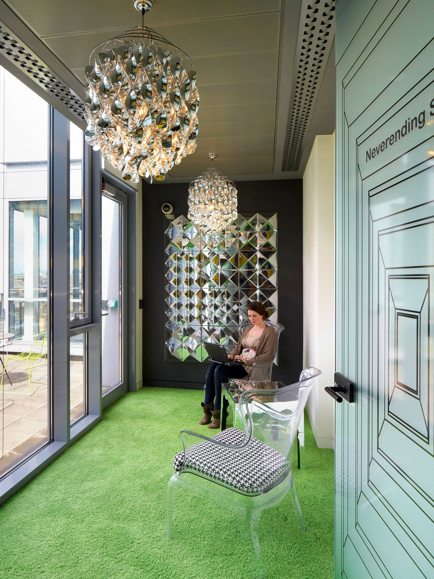 This office space features a vibrant green carpet with a geometric pattern, a large ornate pendant light, and a wall-mounted decorative light piece. A transparent chair with a black and white cushion is positioned by full-height windows, providing ample natural light. A person is seated, using a laptop.