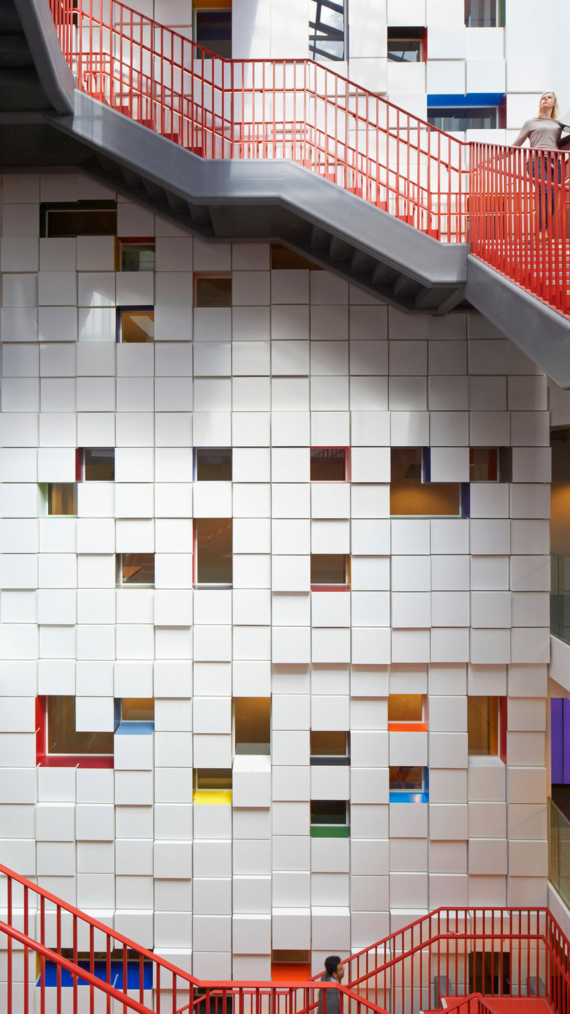 Modern interior featuring a stark white tiled wall with scattered colored transparent panels, complemented by a bold red-hued staircase providing a striking visual contrast.