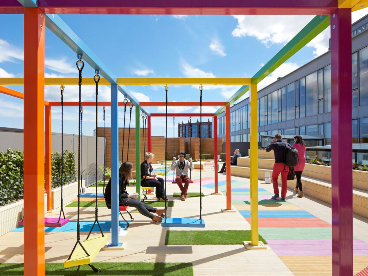 Vibrant rooftop terrace features a playful, multicolored floor design with a series of brightly hued, minimalist frames supporting various swings, fostering a lively social ambiance against a backdrop of clear skies and urban architecture.