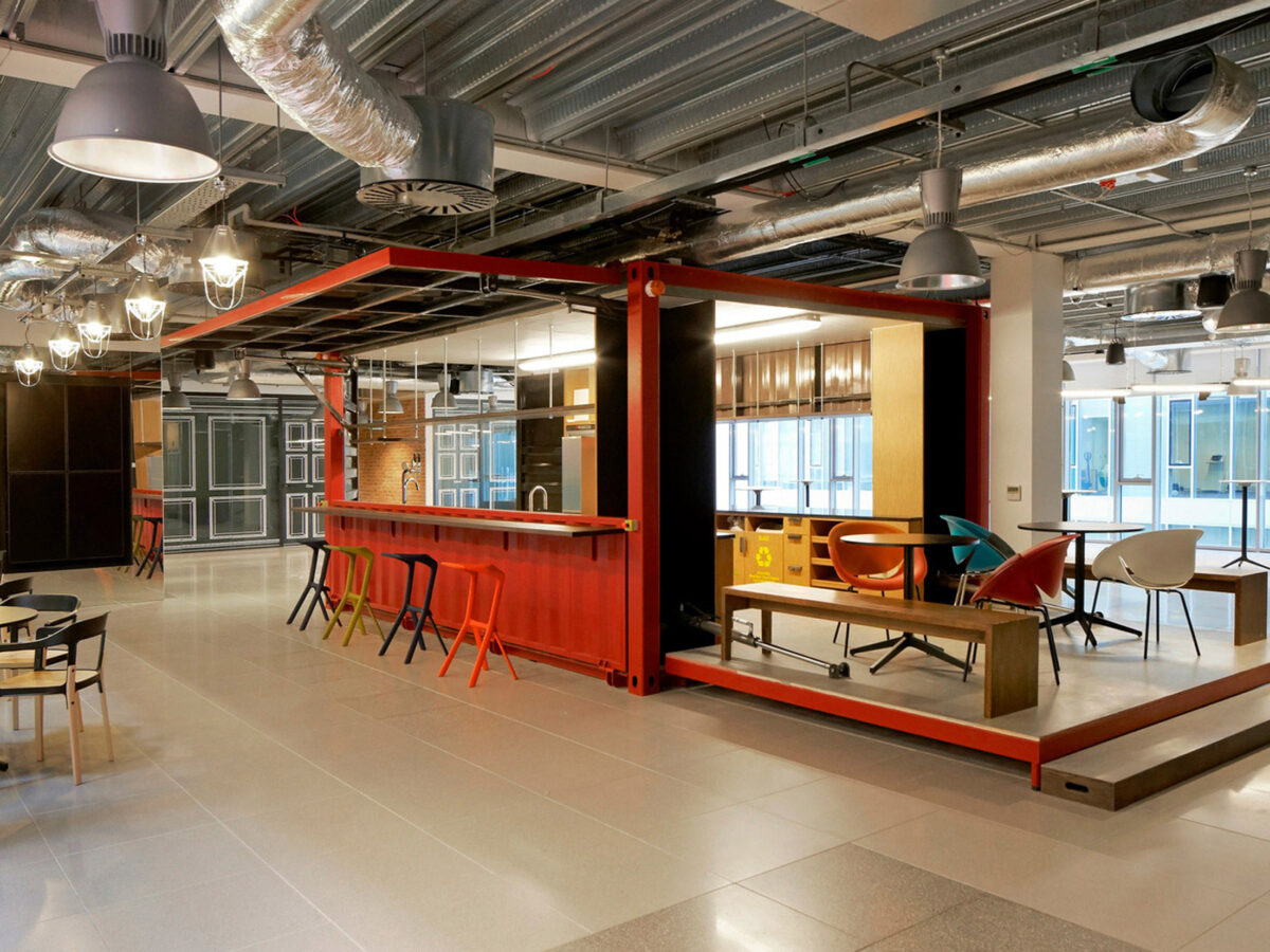 Open-plan office space with exposed ceiling infrastructure, featuring a central, orange-hued kitchenette flanked by eclectic seating arrangements, leading to a vibrant, collaborative work environment. Polished concrete floors reflect strategically placed pendant lighting.