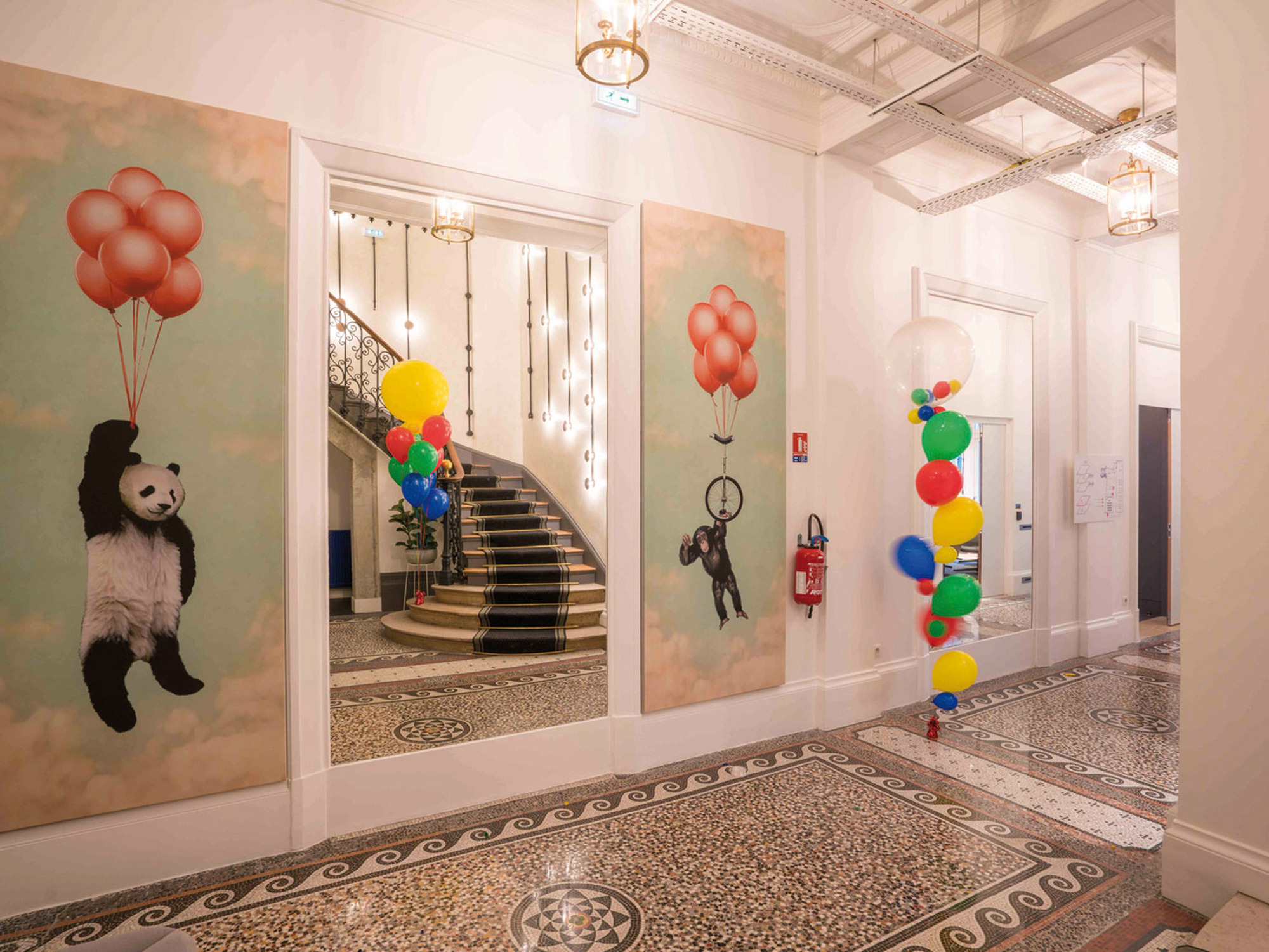 Elegant foyer featuring terrazzo flooring and a grand staircase, accentuated by whimsical artwork of pandas with balloons. Traditional moldings juxtapose modern playful elements, creating an eclectic aesthetic. Vibrant balloon clusters add a three-dimensional pop of color to the space.