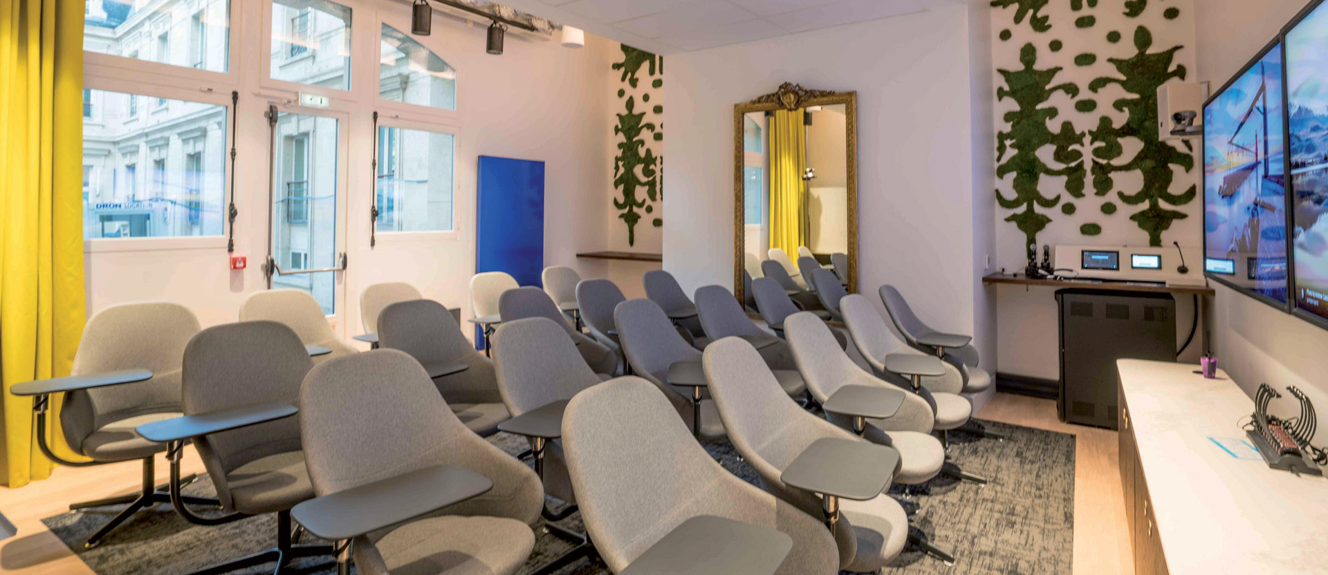 A contemporary meeting room featuring a cascade of ergonomic chairs focused on a large screen; green plant motifs on the walls, yellow accents from a draped curtain, and warm lighting complement the soft gray carpet and white ceiling.