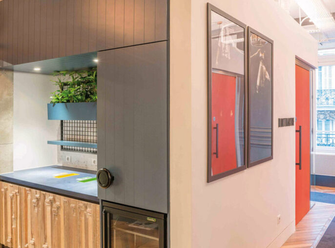 Modern commercial space with an open-concept layout, featuring exposed ductwork, herringbone wood flooring, and a kitchenette with sleek, light wooden cabinetry. Bold geometric patterns and red accents offset the industrial ceiling, creating a dynamic and creative environment.
