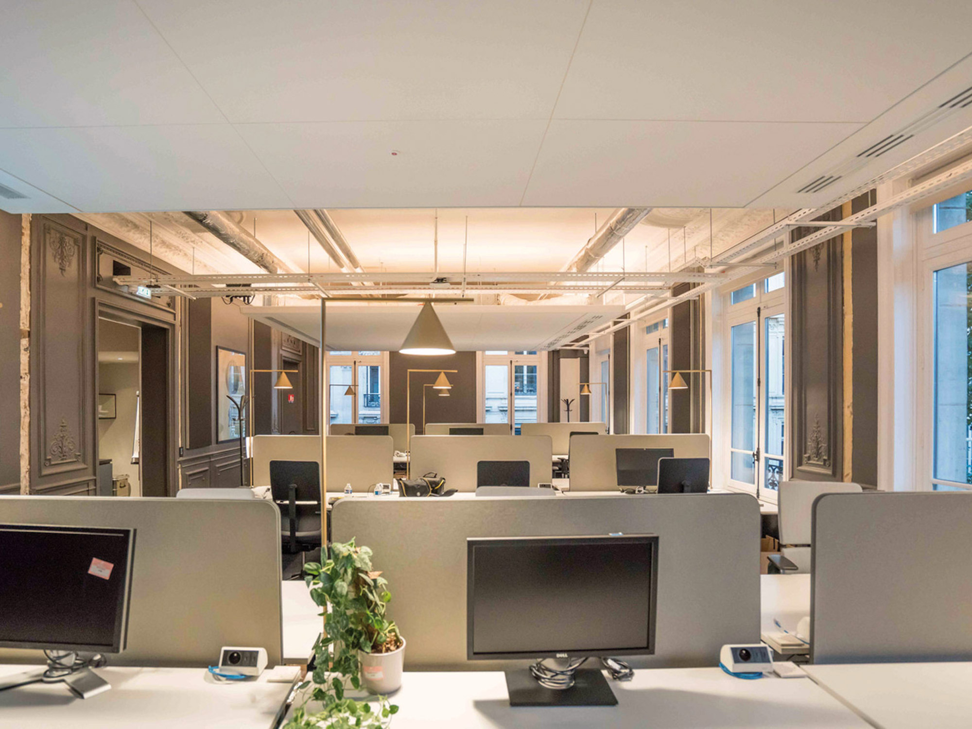 Contemporary office space featuring sleek workstation clusters, pendant lighting, exposed ductwork, and large windows fostering natural light. The color palette is neutral with elegant architectural moldings emphasizing historical charm amidst modern furnishings.