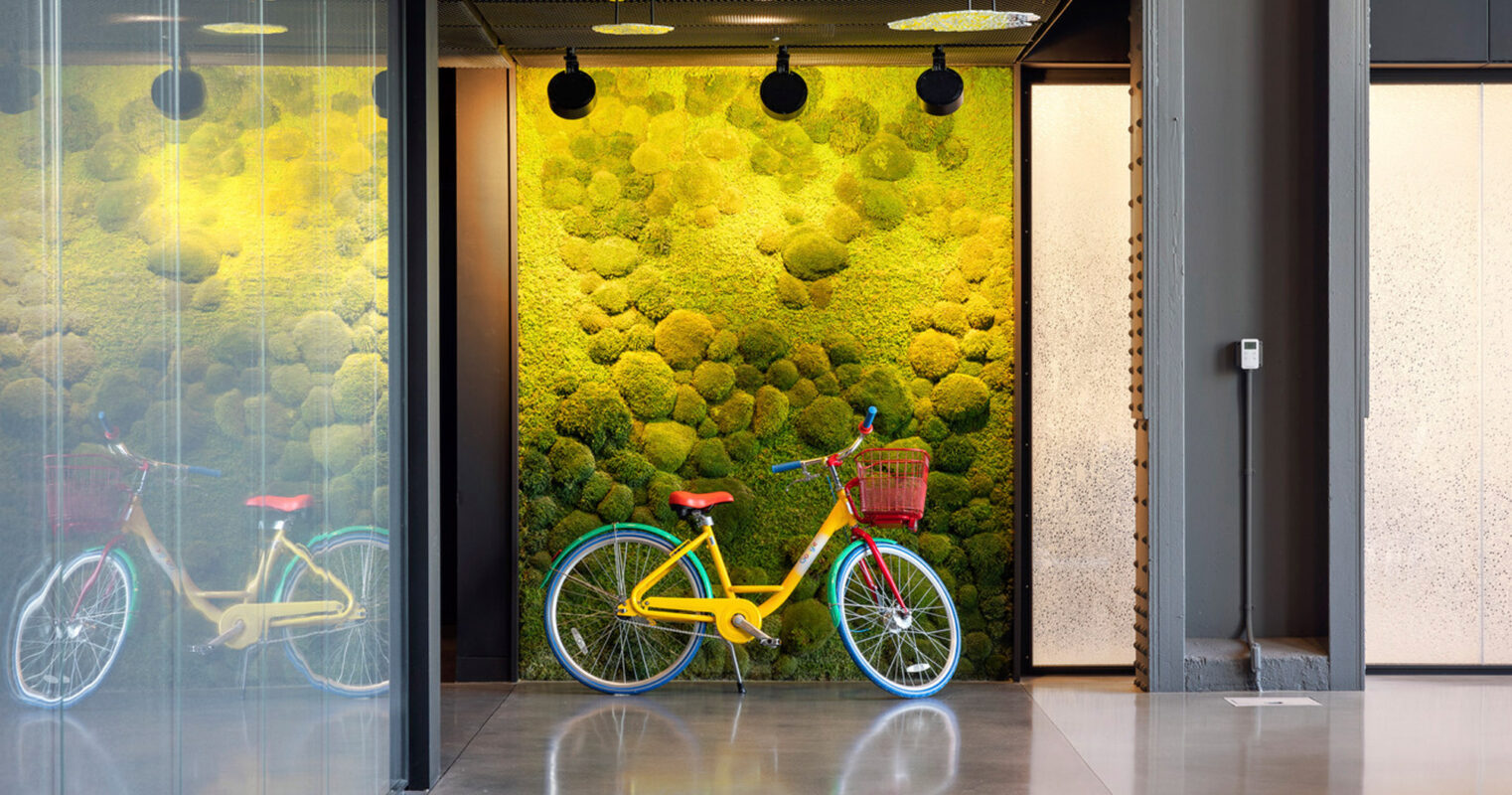 Modern office lobby featuring a vibrant green moss wall, which adds a touch of biophilic design. A bright yellow bicycle with a red basket stands out against the textured backdrop, nestled within a glass-walled niche. Black pendant lights contrast with the industrial gray flooring.
