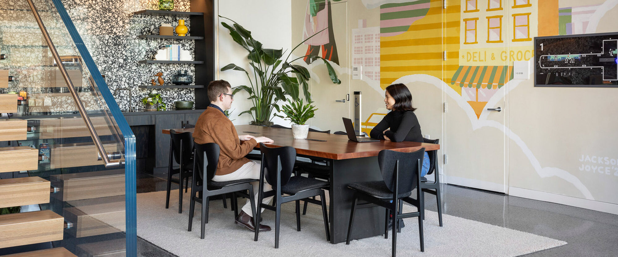 Modern café interior featuring a large vibrant mural with geometric shapes and bicycles, accented by natural wood furniture and a sleek black staircase leading to an elevated area. Two patrons engage in conversation at a central table.