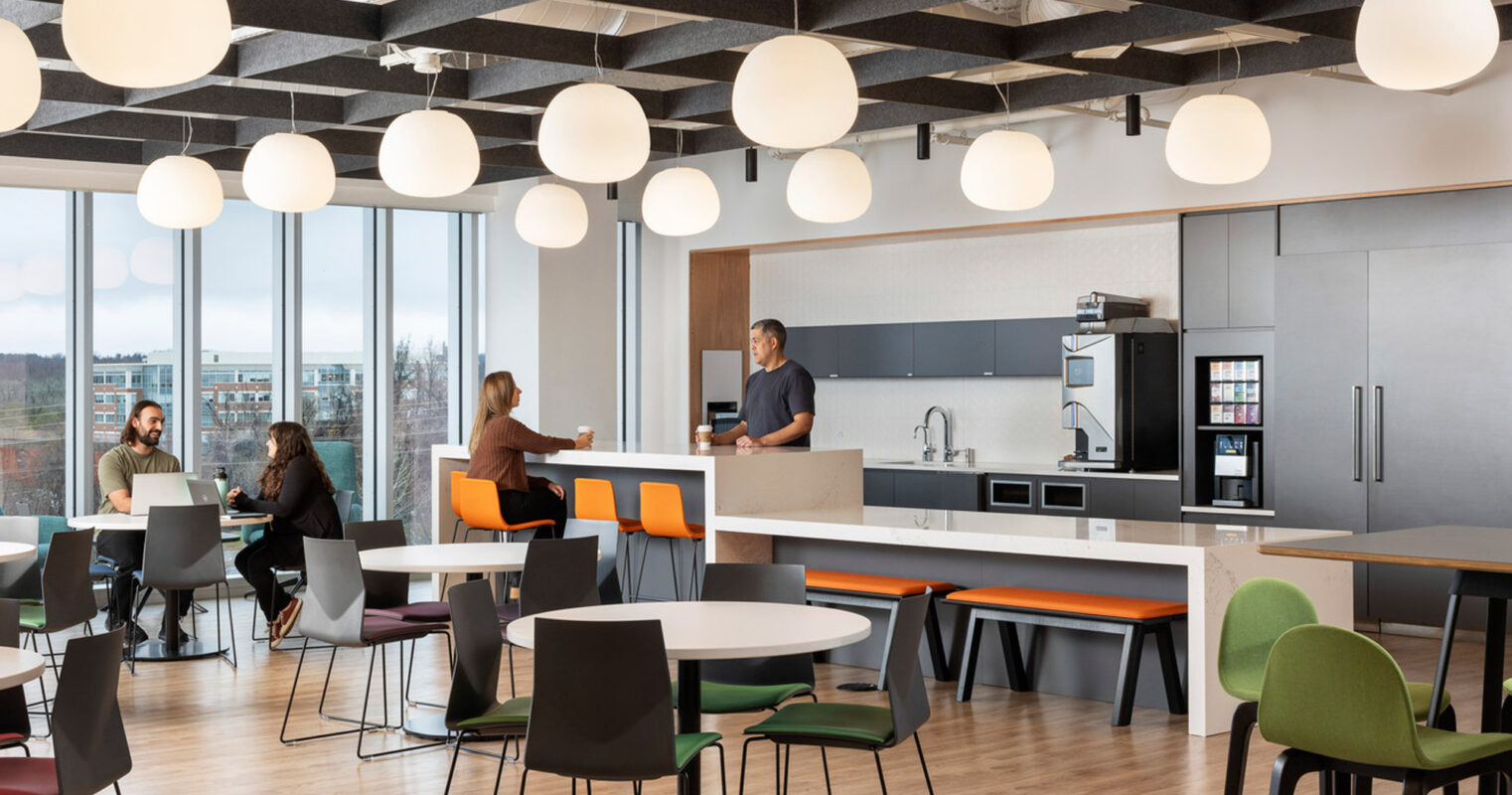 Spacious modern office break room with varied seating arrangements includes a kitchenette, large windows offering natural light, round pendant lights, and a combination of vibrant and neutral color palette enhancing the welcoming ambiance.